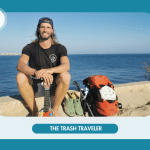 Mann am Strand mit "The Trash Traveler"-Logo auf Rucksack und T-Shirt, Gitarre in der Hand.