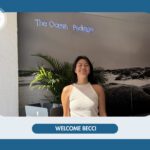 Becci, die neue Praktikantin bei The Ocean Package, lächelnd im Büro vor einer Wand mit Logo und Pflanzen.
