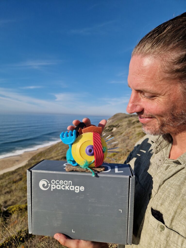The Trash Traveler hält die Trash Art Awards Trophäe und eine Ocean Package Mehrwegbox am Strand, als Beispiel für zirkuläre Wirtschaft und systemischen Wandel.