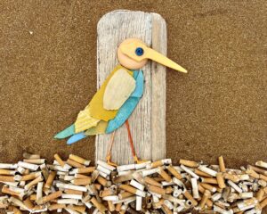 Vogel-Collage auf Treibholz, bestehend aus Plastikfragmenten und umgeben von Zigarettenkippen, als Beispiel für die kreative Wiederverwendung von gefundenem Müll.