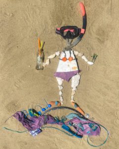 Taucherfigur aus Muscheln, Plastik und Tauchausrüstung auf Sand, die Müll als Ressource für Upcycling-Kunst zeigt.
