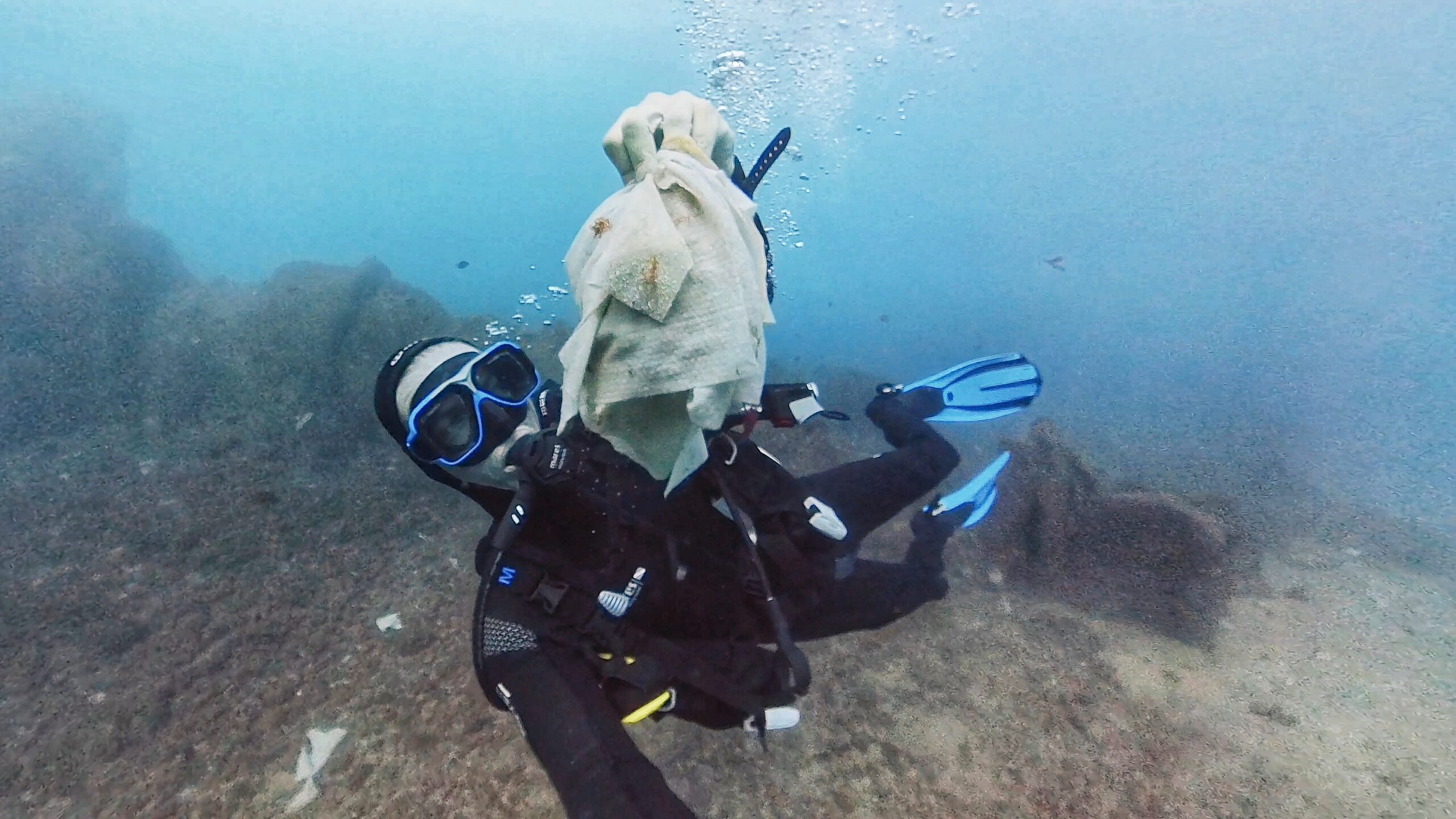 Taucher schwimmt mit einer großen Menge Feuchttücher über einer felsigen Unterwasserlandschaft, symbolisiert Meeresverschmutzung.