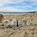 Nahaufnahme von weggeworfenem Plastikmüll und medizinischen Abfällen, die senkrecht im Sand eines Strandes stecken.