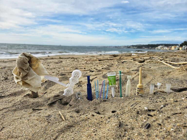 Nahaufnahme von weggeworfenem Plastikmüll und medizinischen Abfällen, die senkrecht im Sand eines Strandes stecken.