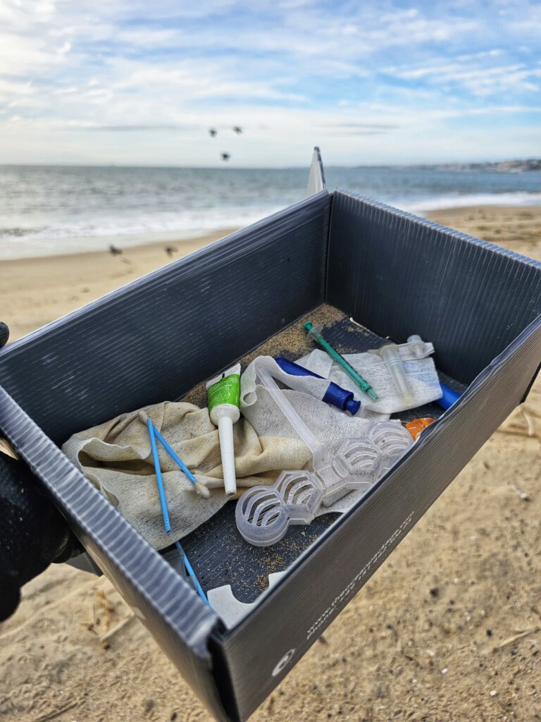Eine Hand hält eine graue, robuste Mehrweg-Versandbox von The Ocean Package, in der gesammelter Plastikmüll vom Strand liegt. Im Hintergrund ist das Meer zu sehen.