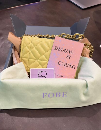 Eine Mehrweg Versanbox steht geöffnet auf dem Boden. Der Inhalt zeigt eine gelbe Handtasche, eine Karte mit der Aufschrift “sharing ist caring” und ein Beutel für die Tasche mit dem Logo von FOBE.